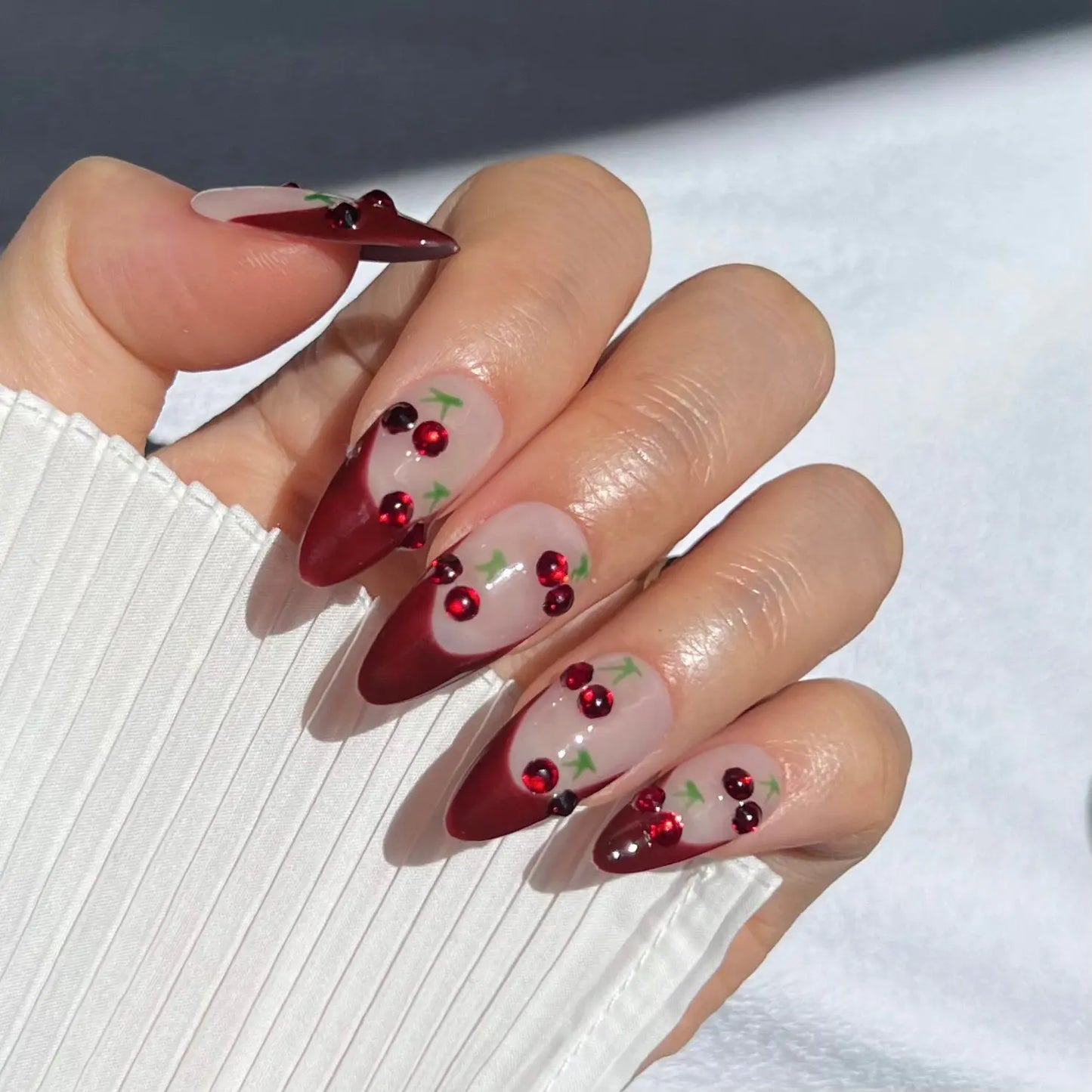 A close-up of a hand wearing almond-shaped press-on nails with red tips and cherry designs, offering a fun and vibrant look.