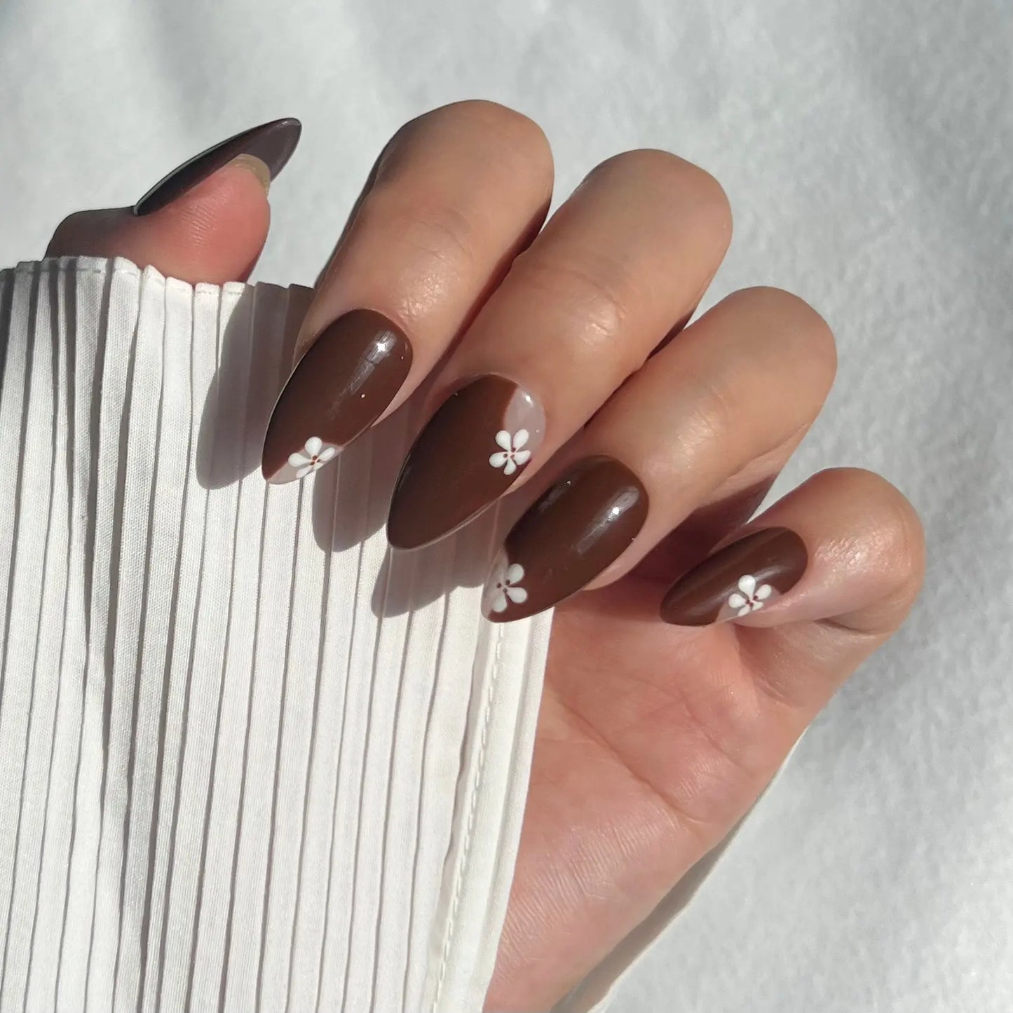 A close-up of a hand wearing almond-shaped press-on nails with a glossy, earthy tone and a touch of shimmer, creating a chic yet subtle look.