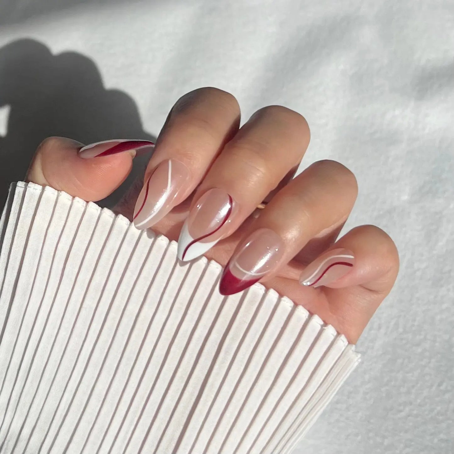 A close-up of a hand wearing almond-shaped press-on nails with burgundy and white line designs on a nude base, offering a chic and modern style.