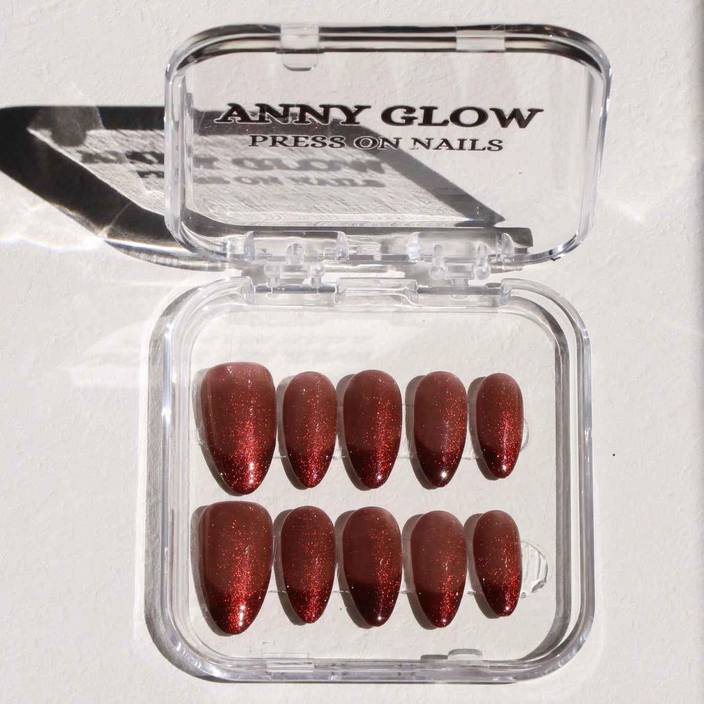 A set of press-on nails displayed in a clear plastic box, featuring a deep red hue with shimmering glitter, creating a bold and radiant look.