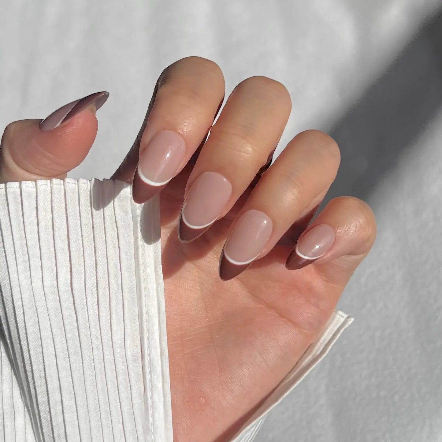 A close-up of a hand wearing almond-shaped press-on nails with a nude base, brown tips, and a white accent line, offering a modern, elegant appearance.
