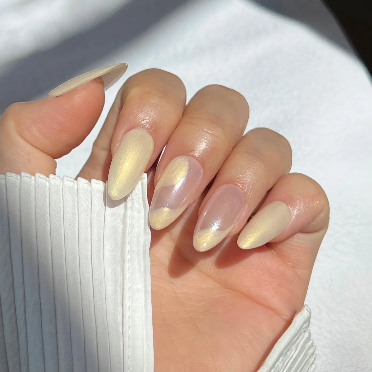 A close-up of a hand wearing almond-shaped press-on nails with a pearlescent finish, showcasing soft, dreamy colors and wavy patterns for an ethereal look.