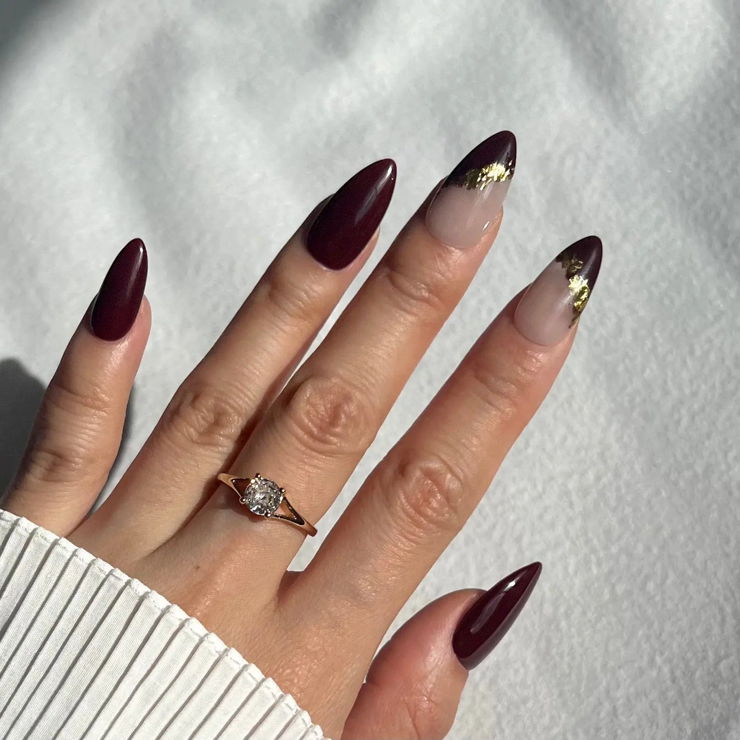 A set of press-on nails with a deep burgundy base and translucent tips adorned with shimmering gold leaf accents. The nails are neatly arranged in a clear plastic case.