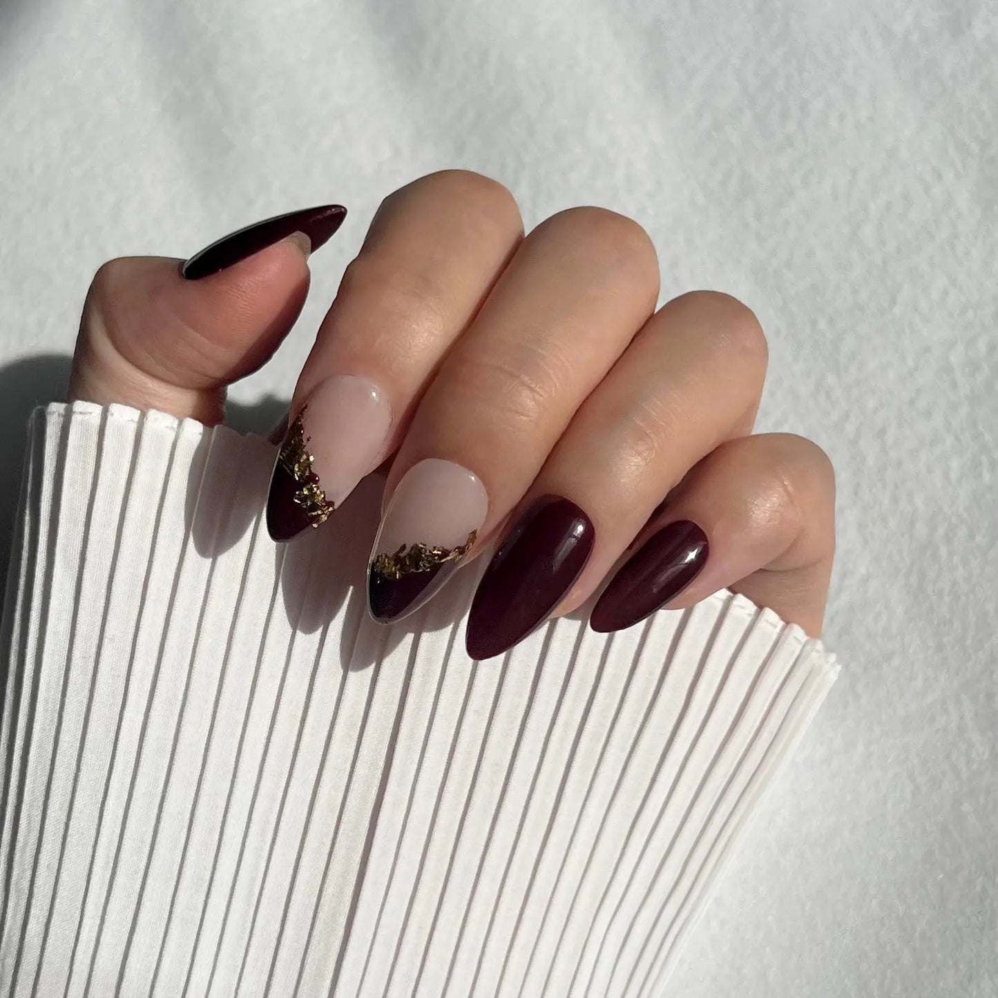Close-up of a hand wearing press-on nails with a rich burgundy color and translucent tips with gold leaf accents, creating a regal and sophisticated look.