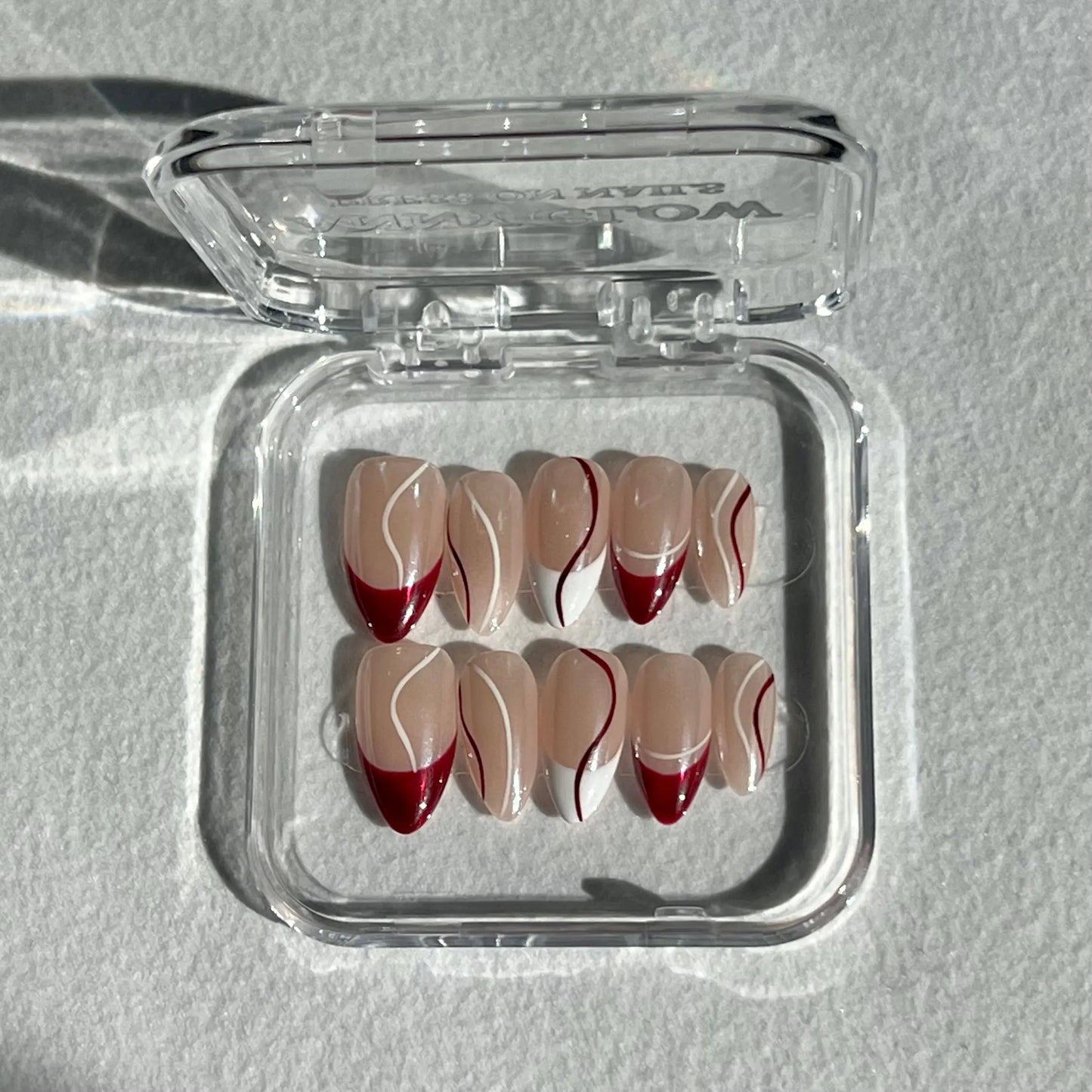 A set of press-on nails displayed in a clear plastic box, featuring a nude base with elegant burgundy and white line designs for a refined, everyday look.