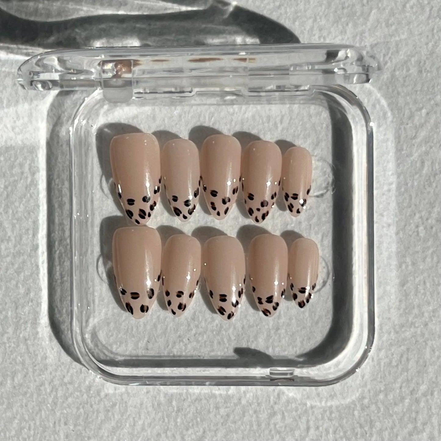 A set of press-on nails displayed in a clear plastic box, featuring a nude base with black leopard print accents for a chic and playful design.
