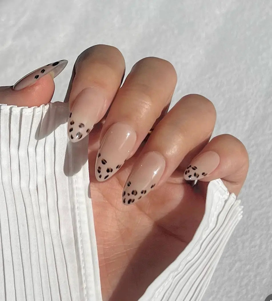 A close-up of a hand wearing almond-shaped press-on nails with a nude base and black leopard print accents, creating a bold and trendy look.
