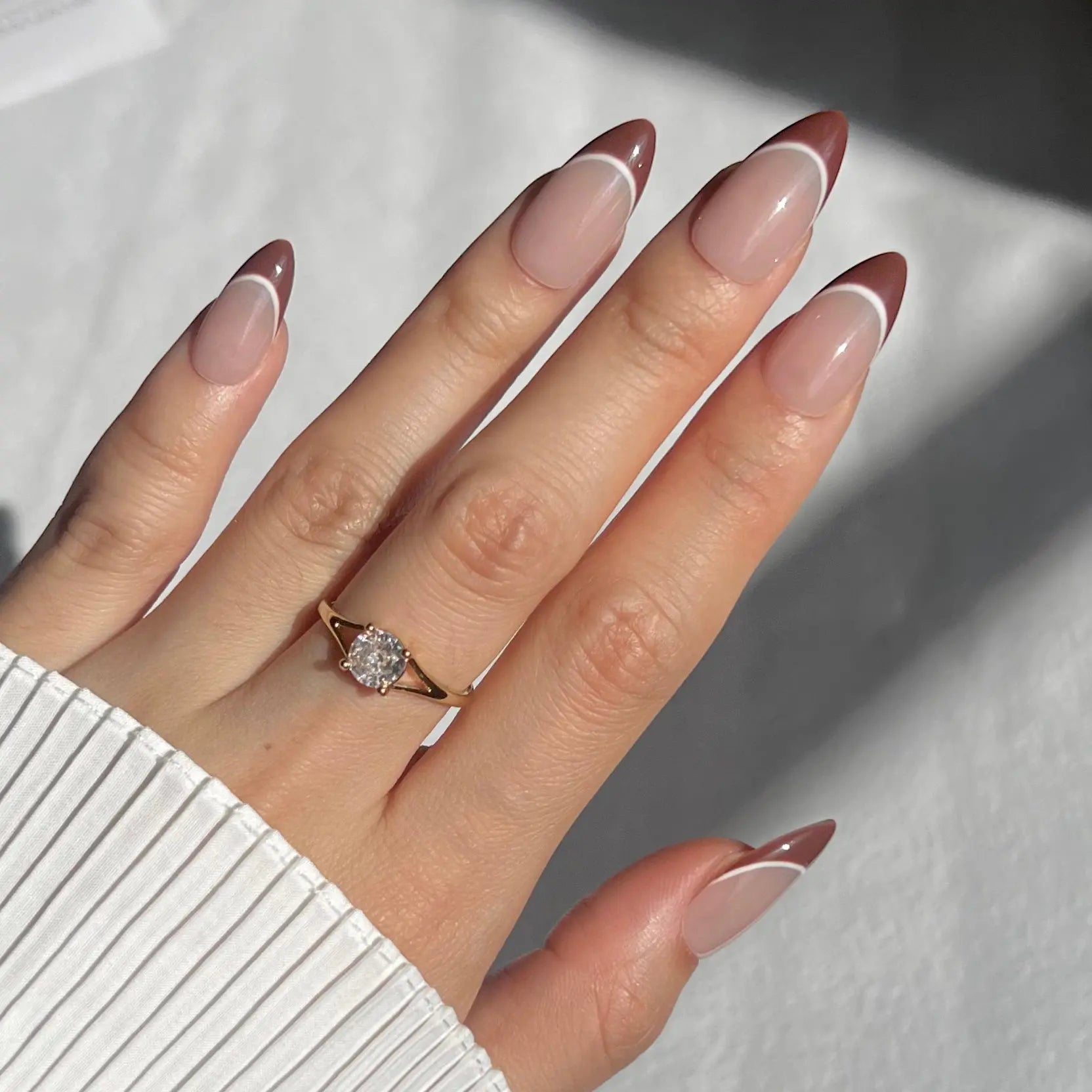 A hand showcasing press-on nails with brown tips and white lines over a nude base, perfect for a sleek, refined style